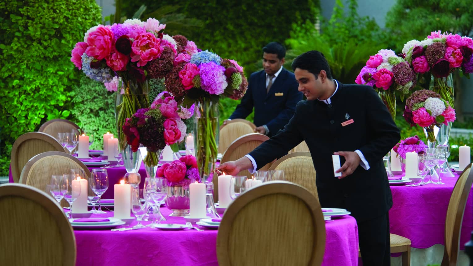 Two male staff set table with pink tablecloth, large vases of flowers and candles