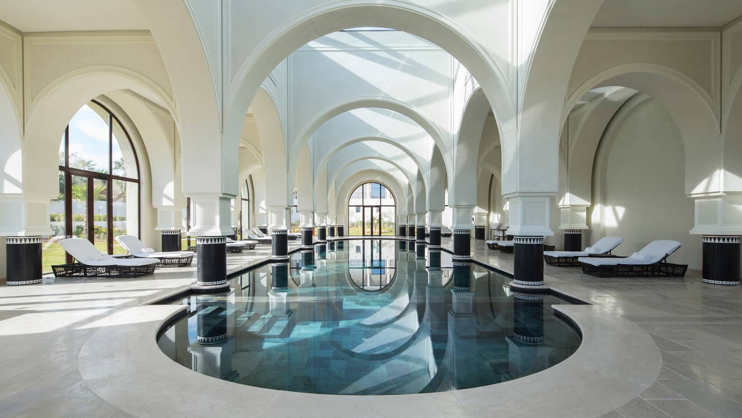 Oval indoor swimming pool under soaring, arched white pillars, windows