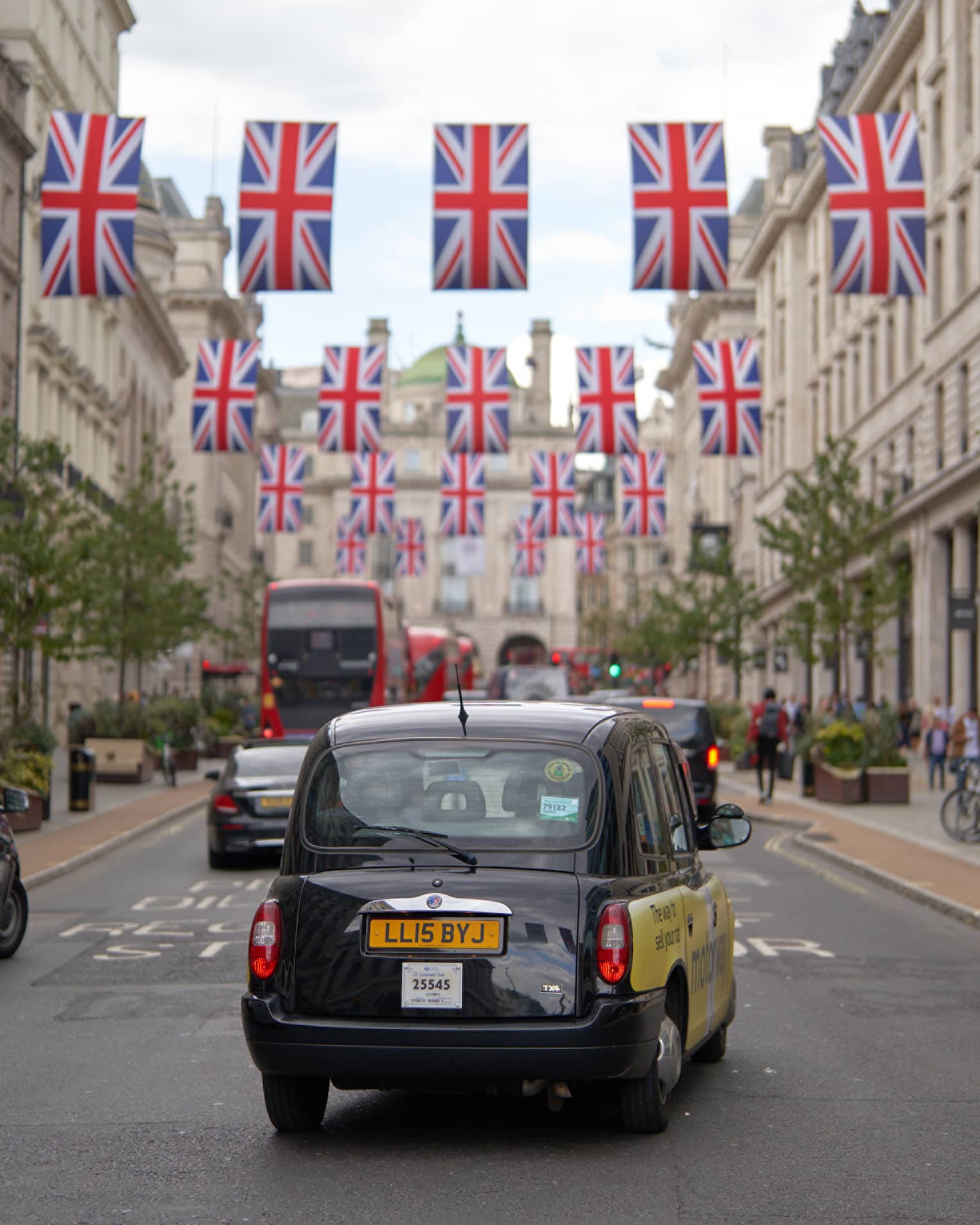 Rear view of London-style black cab, double-decker buses and other cars on road strung across with rows of Union Jack flags.