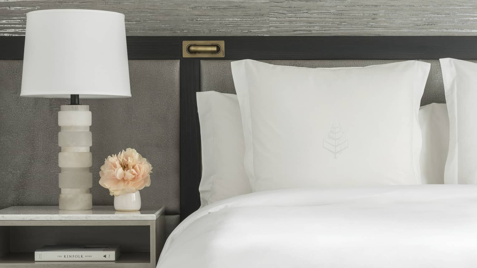 Luxury hotel room king bed with white sheets and Four Seasons tree logo embroidered on sham, next to a nightstand with white lamp and small vase of peach-coloured flowers