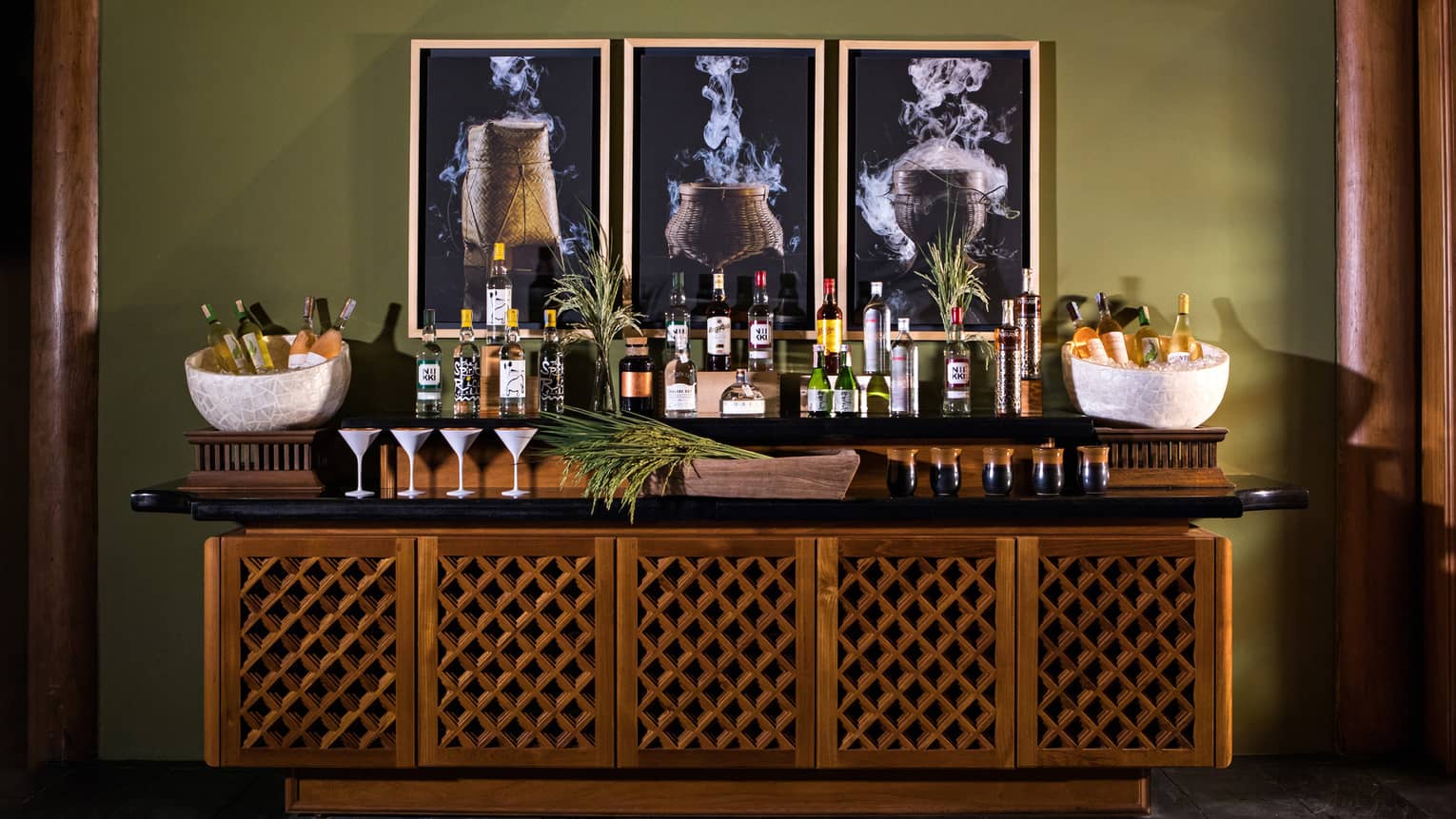 Wood buffet with liquor bottles, martini glasses, wine on ice in bowls, three framed prints