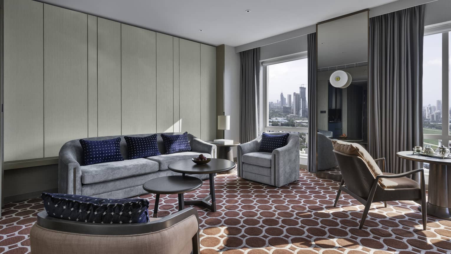 Renovated Executive Suite living area with grey sofa and armchair, round nesting coffee table, floor-to-ceiling windows with city views, red patterned carpet