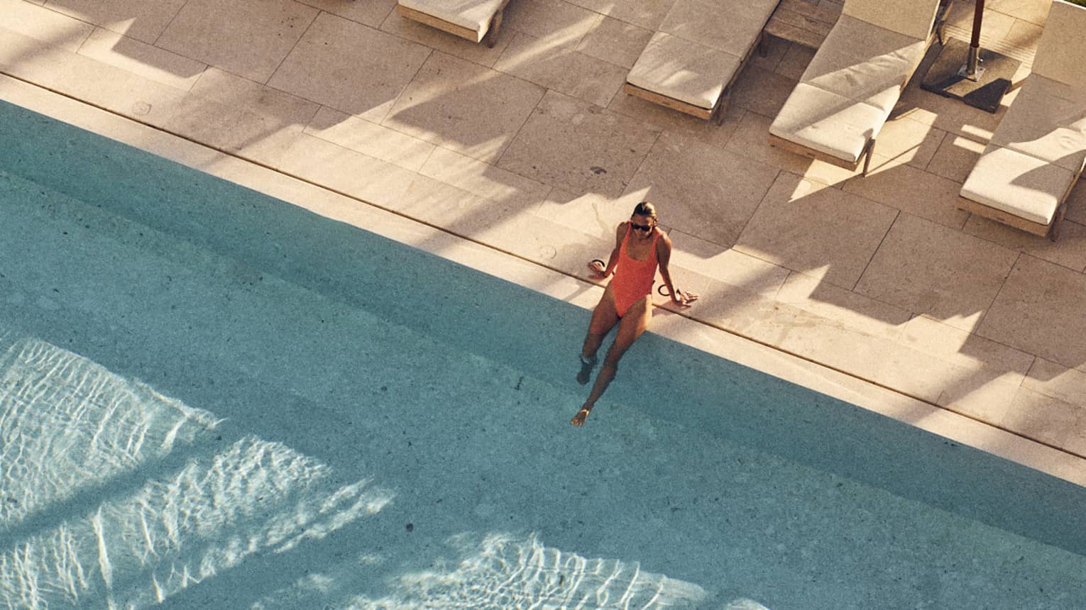 Aerial view of a woman in an orange swimsuit sitting on the edge of a large pool surrounded by lounge chairs and white umbrellas