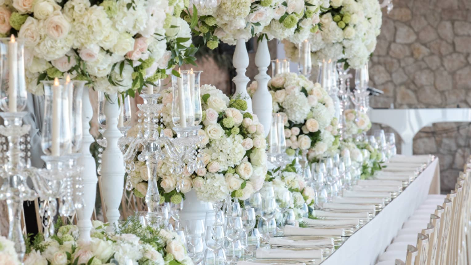 Elegantly set white banquet table lined with chairs, tall white and pink floral arrangements, candles