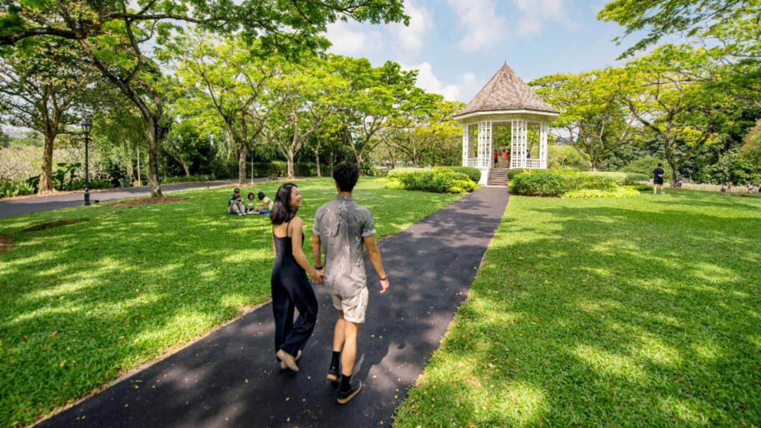 A man and woman hold hands and walk along an asphalt trail through the Singapore Botanic Gardens