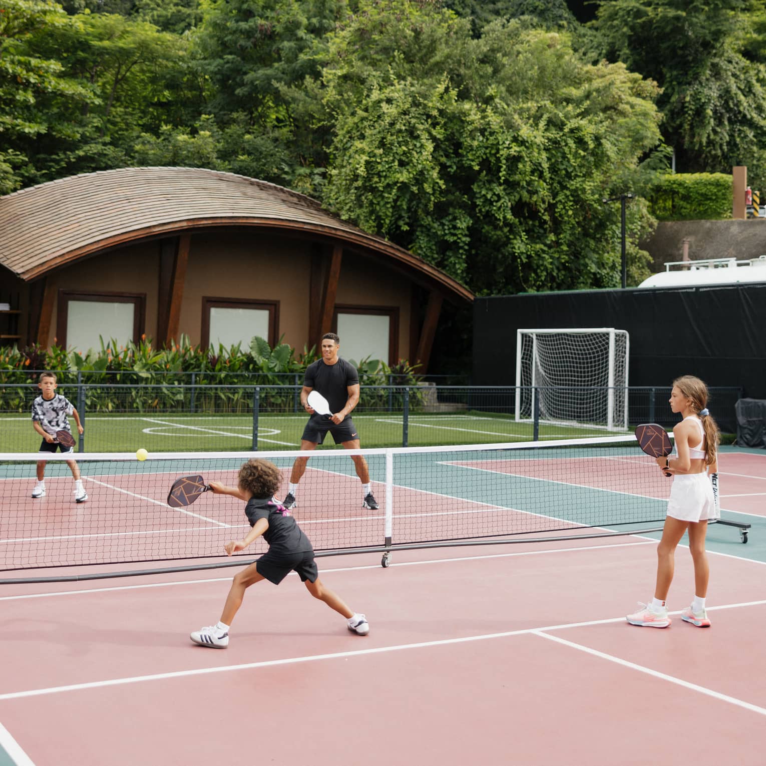 One adult and three children play pickleball on an outdoor court surrounded by trees