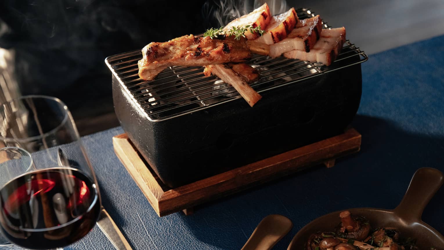 Sliced meat atop small tabletop grill with glass of red wine and two side dishes in small pans