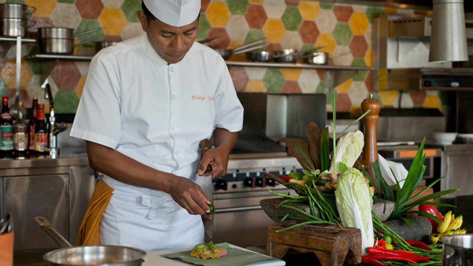Chef in white uniform squeezes lime on large kitchen knife over dish, next to heads of fresh lettuce and whole peppers