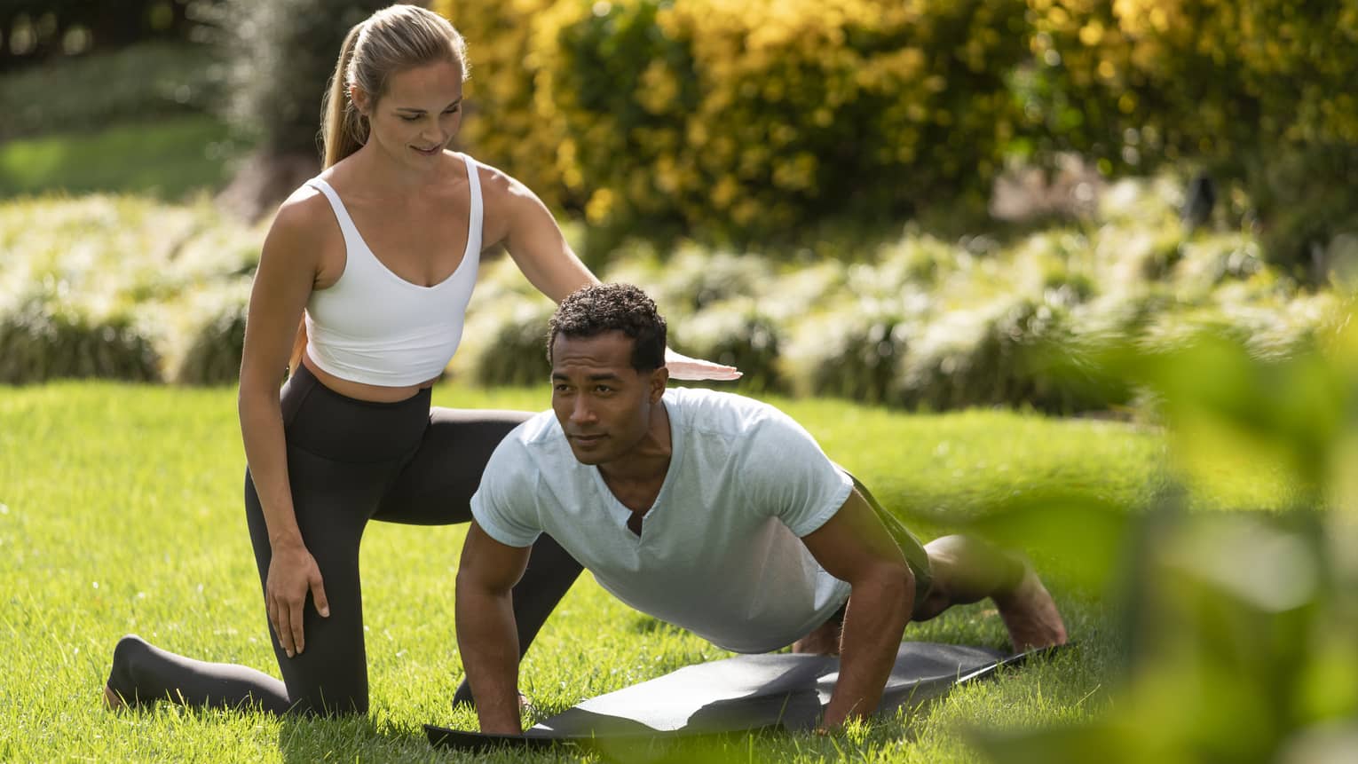 A yoga instructor helping a man get into a yoga position outside on very green grass.