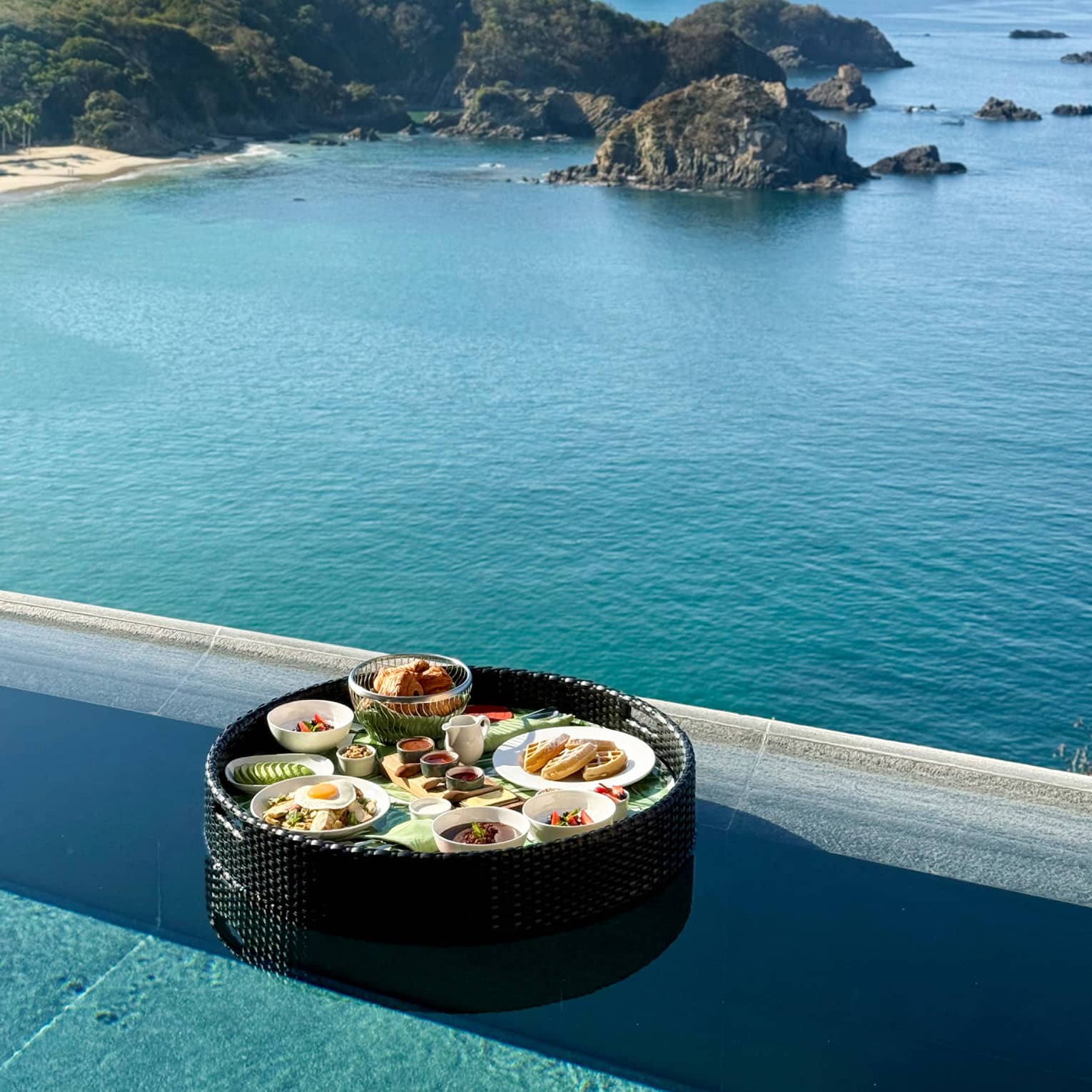 A tray of plated breakfast food items on a tray floating near the edge of an infinity pool, with coastline and ocean views.