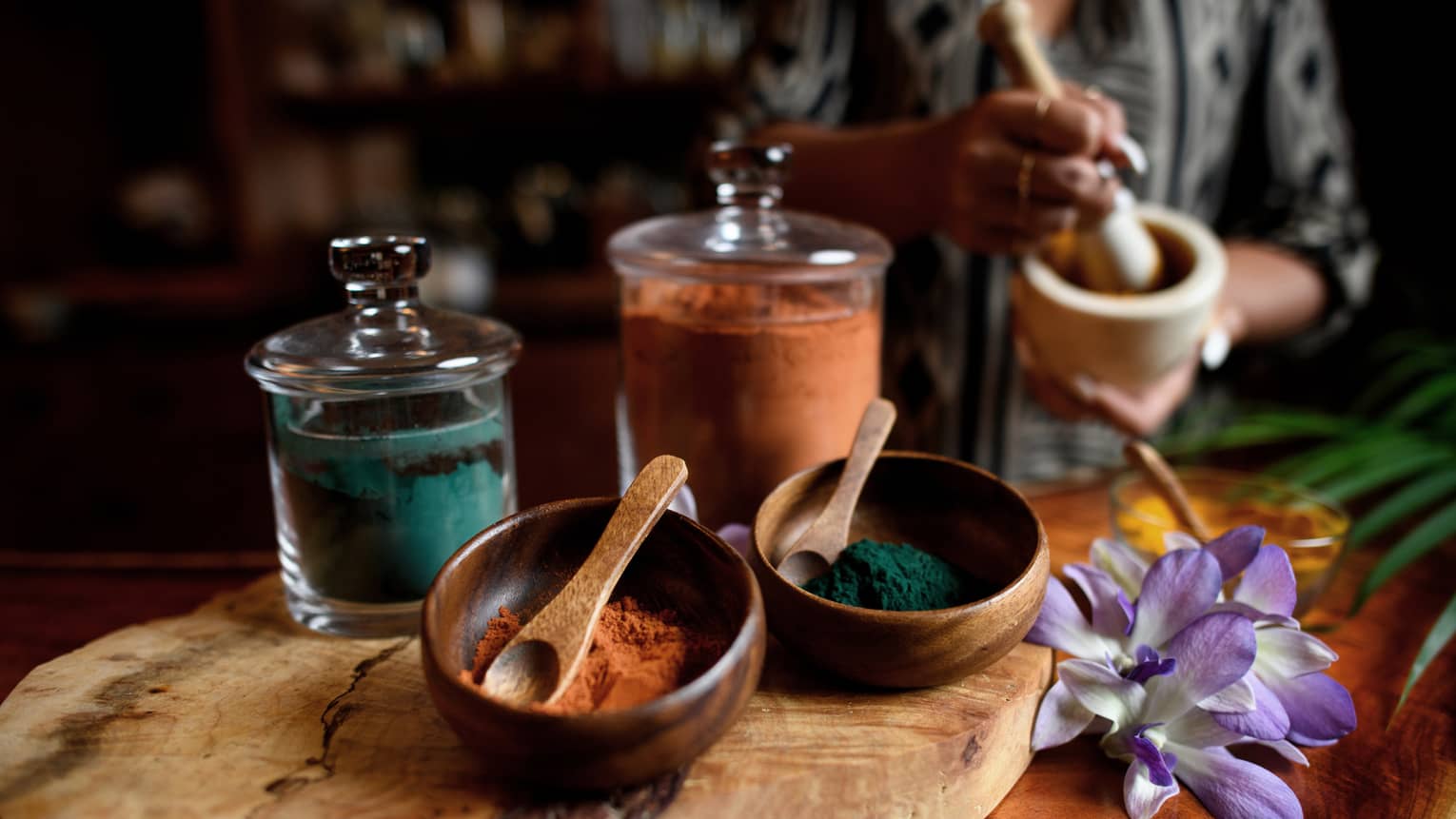 Small wood bowls and spoons beside glass jars filled with colourful powders, tropical flower