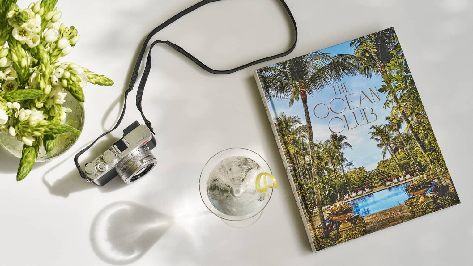 The Ocean Club Assouline book on a white surface next to a martini glass with a lemon twist garnish and camera