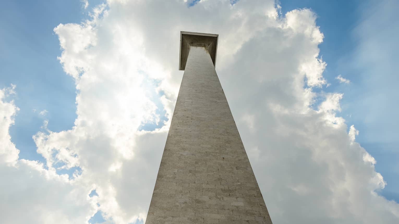 Staring up from the base of the Monas National Monument; a blue sky and the sun peeking through the clouds as a backdrop.