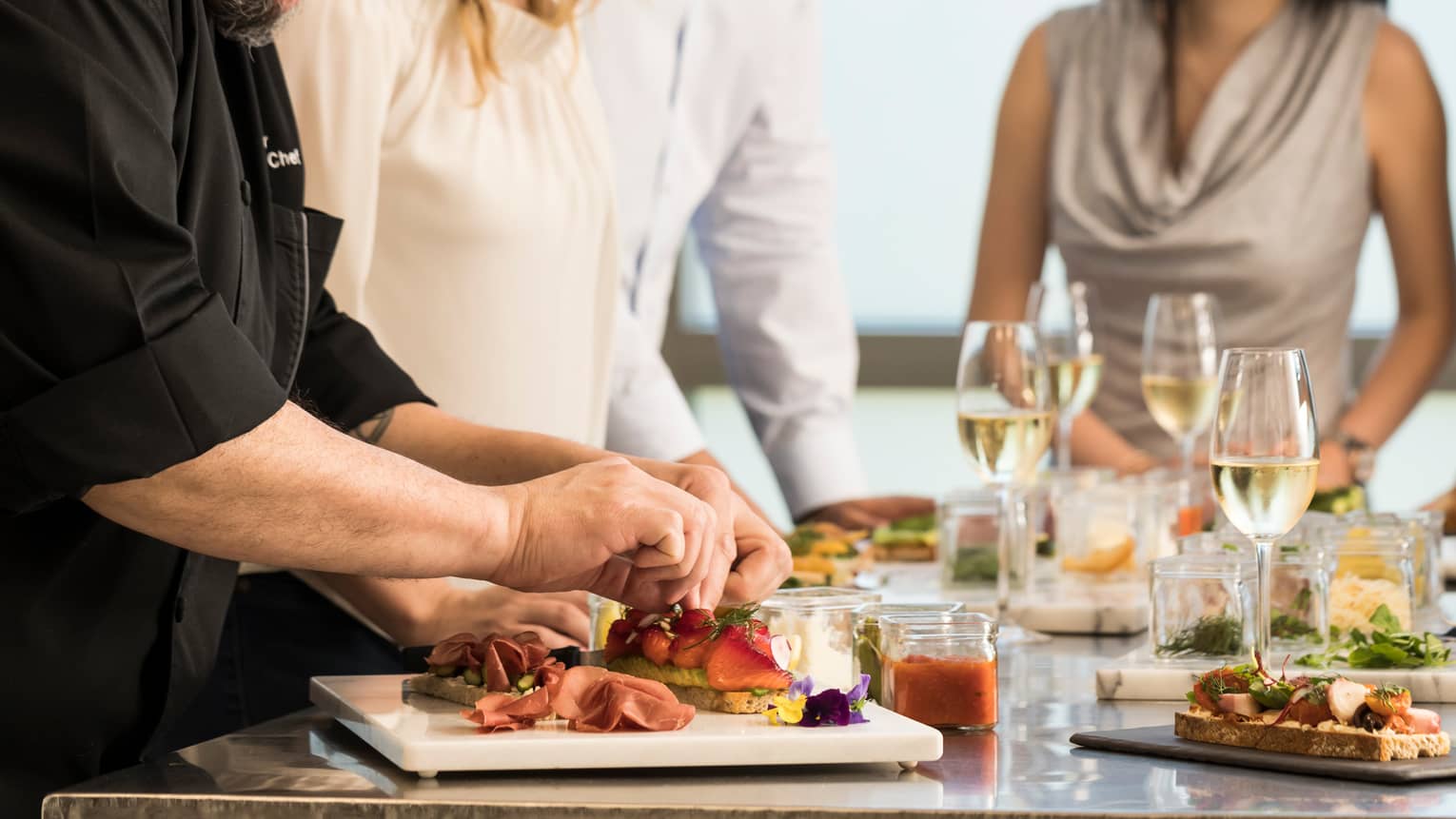 Chef assembles food at counter with smiling group of friends, white wine in glasses