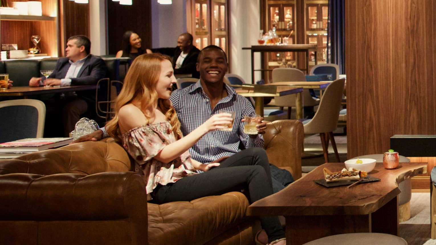 A man and woman sitting on a couch in a bar area drinking and laughing.