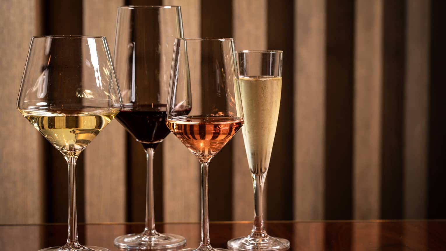 Four modern wine glasses holding white, red and rose wine are lined up on a wooden table 