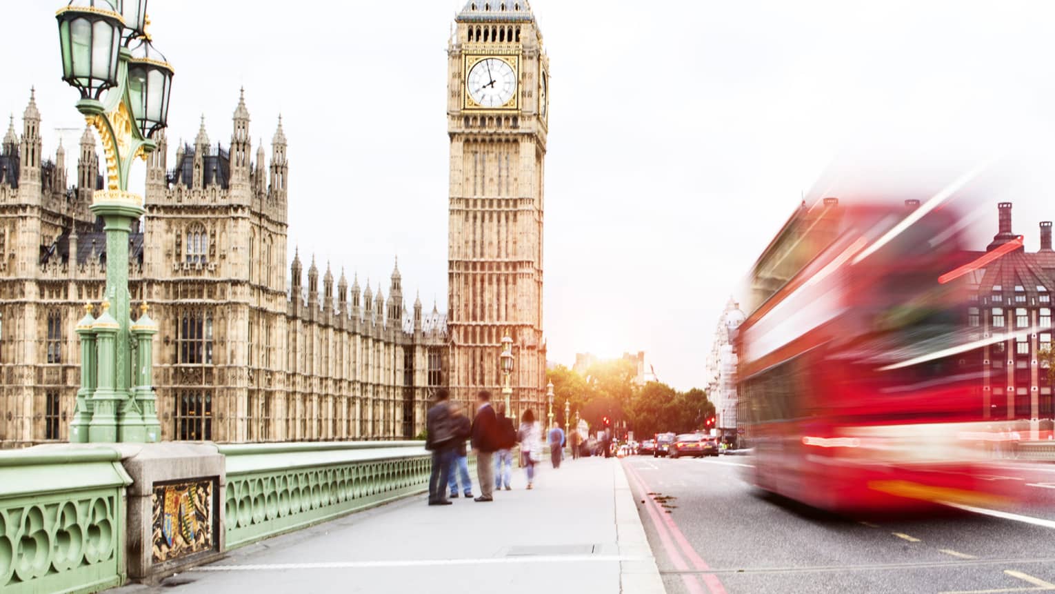 Red double-decker bus speeds by, Big Ben in the background as tourists stand nearby 
