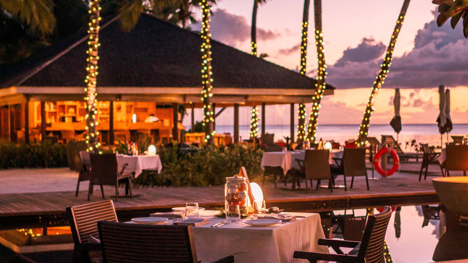 Claudine waterfront terrace with square dining tables amid palm trees wrapped in white lights at dusk