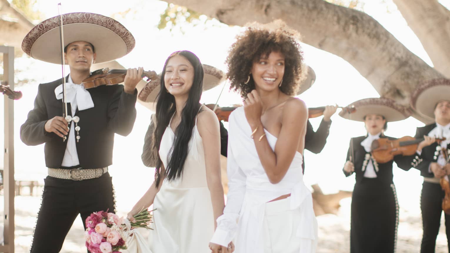 A wedding photo of two brides holding hands and walking down a sandy beach to the alter, there are men and women in black clothing playing violins around them.