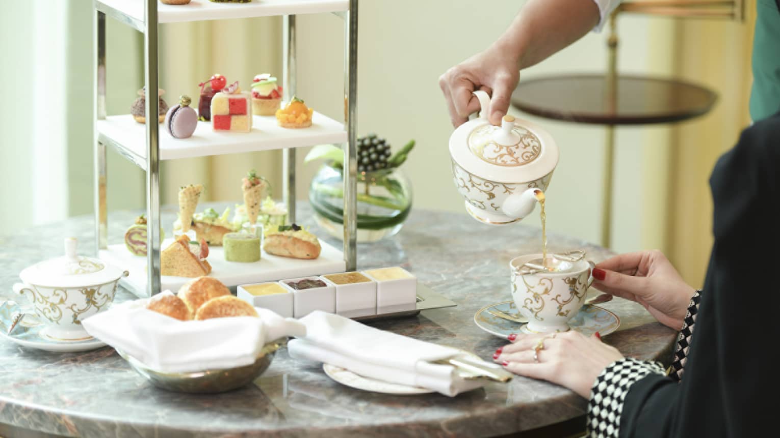 A shot of a man's hand pouring a cup of tea for a woman, with a large dessert platter in the background.
