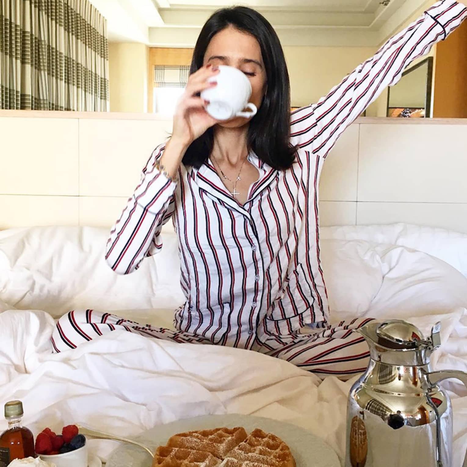 Woman on bed sips coffee cup, stretches in front of in-room breakfast dining tray