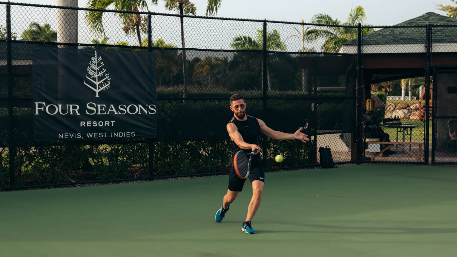 A person playing tennis on a tennis court, with palm trees and blue sky in the background
