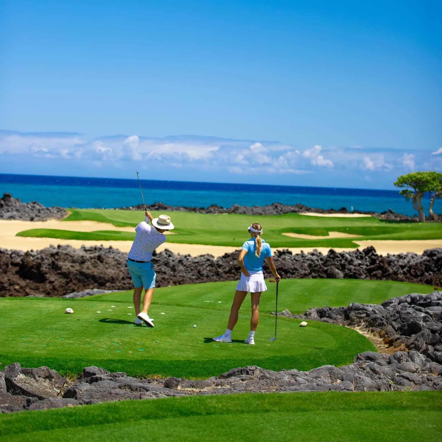 Two people playing golf on green fairways with black lava rock and the ocean in the background.