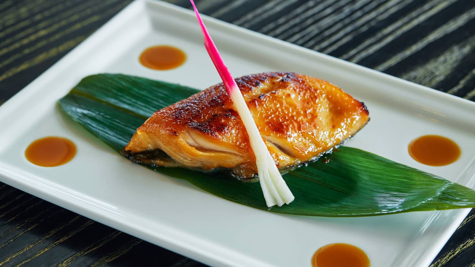 Black Cod Yuzu Miso fish fillet marinated, oven-baked on grilled seafood filet on leaf on white platter