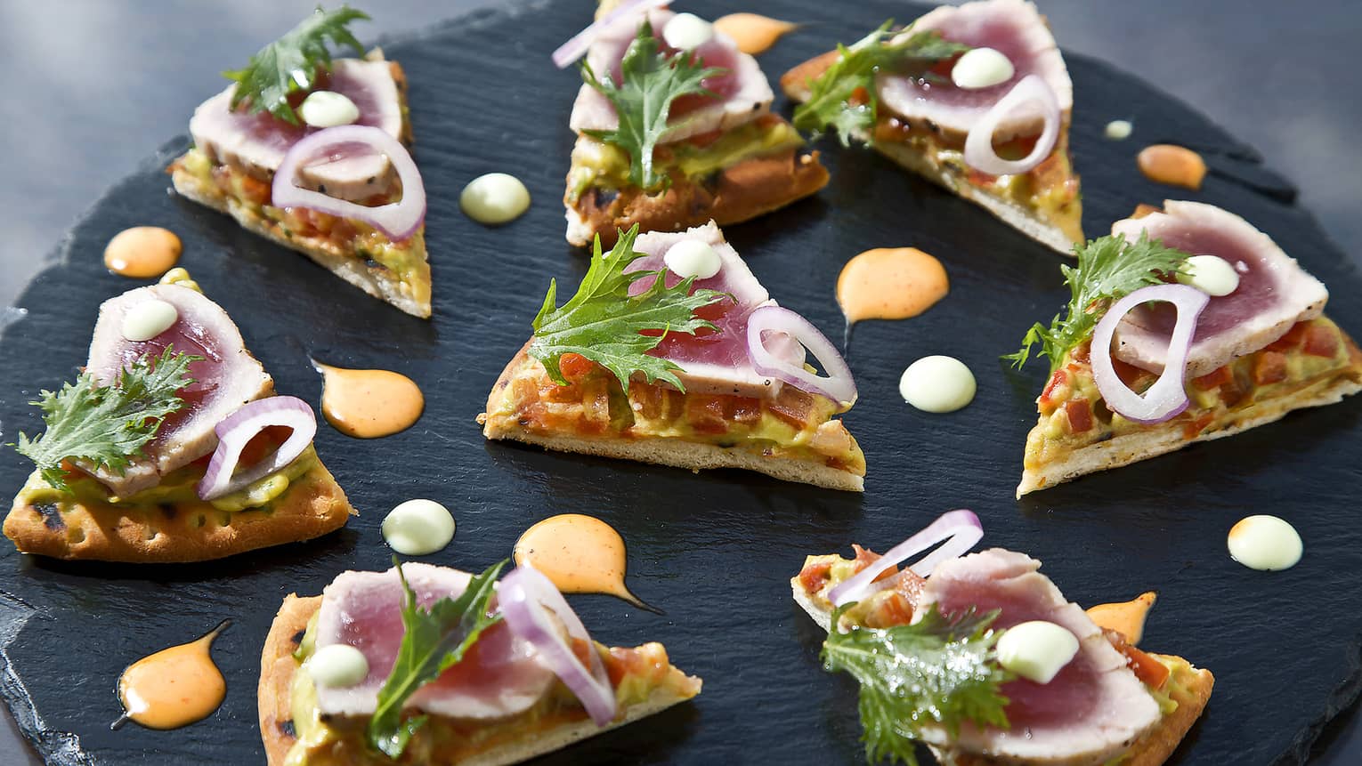 Small gourmet flatbread slices topped with red onion, greens on slate platter