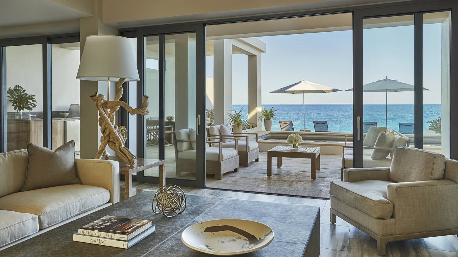 Elegant living room with neutral-toned seating, a coffee table and sliding glass doors opening to a terrace with ocean views and shaded loungers
