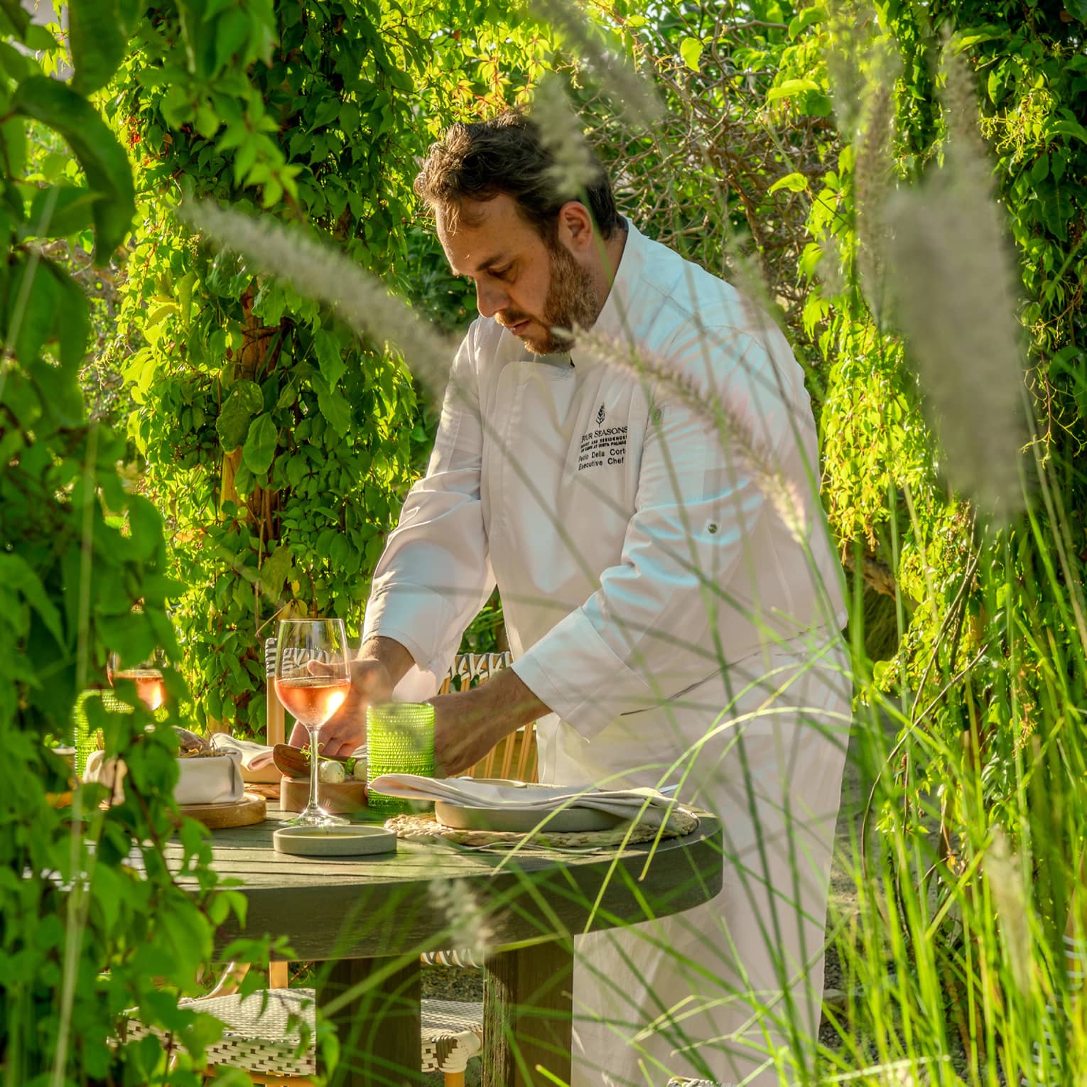 A chef works at a small dining table in a lush, green garden setting.