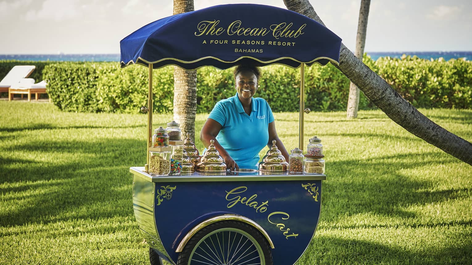 Smiling woman standing behind a blue gelato cart by The Ocean Club, Four Seasons Resort Bahamas, with a green lawn and ocean view in the background