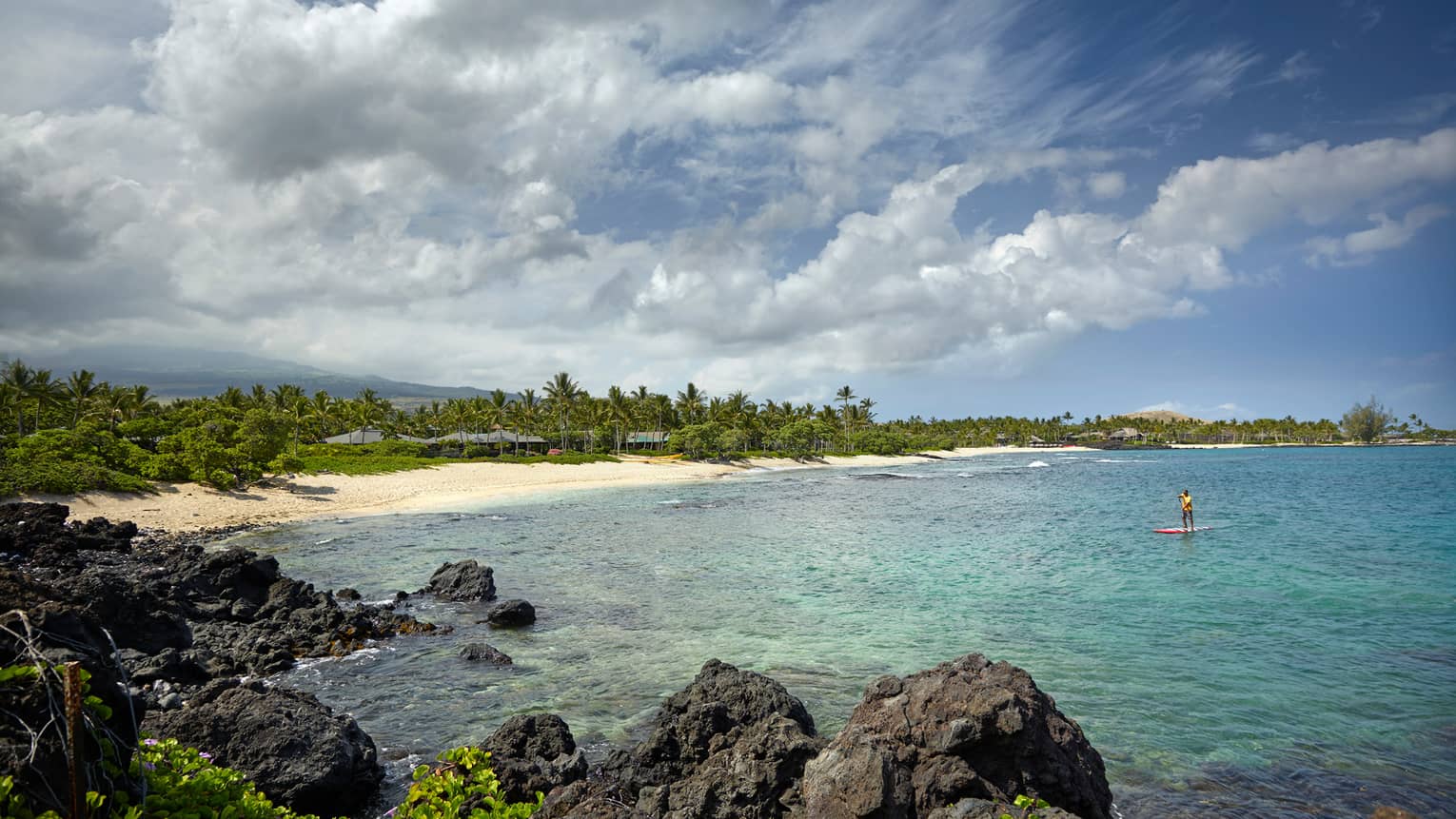 Person on stand-up paddleboard in lagoon past large black lava rocks, near white sand beach