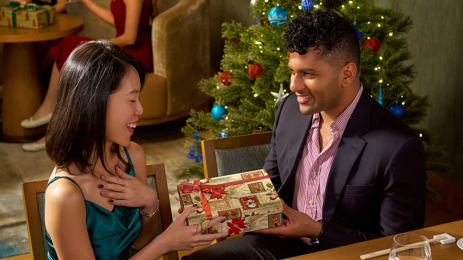 Two guests sitting at a dining table in a restaurant, one person giving the other person a gift.