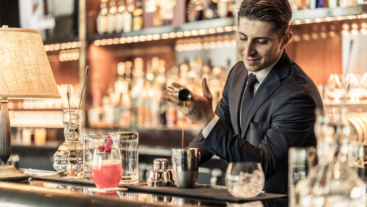 Bartender behind bar, pouring liquid from jigger into metal shaker among bar accessories
