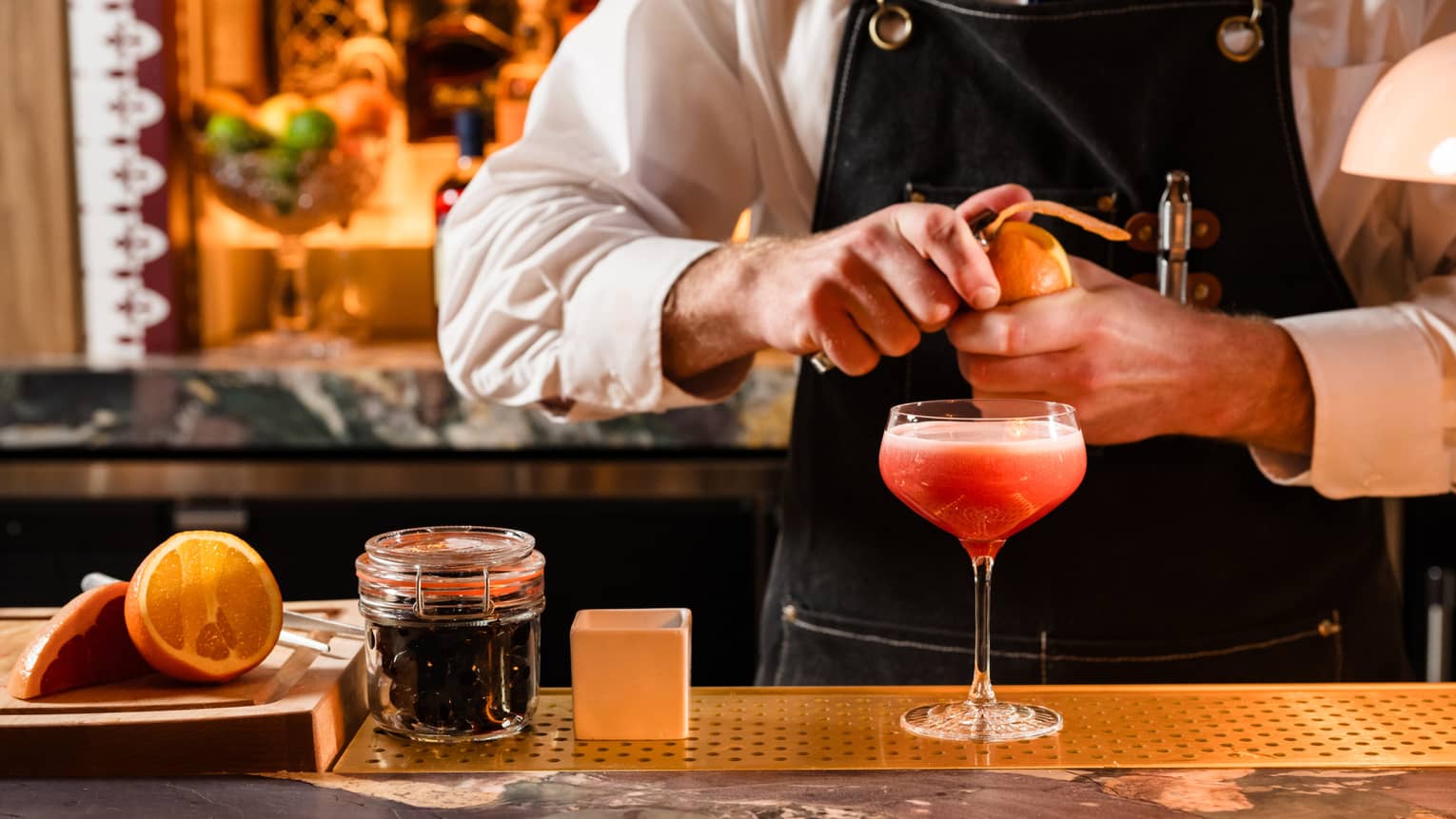 A bartender prepares a reddish drink in a stemmed glass on a bar counter.