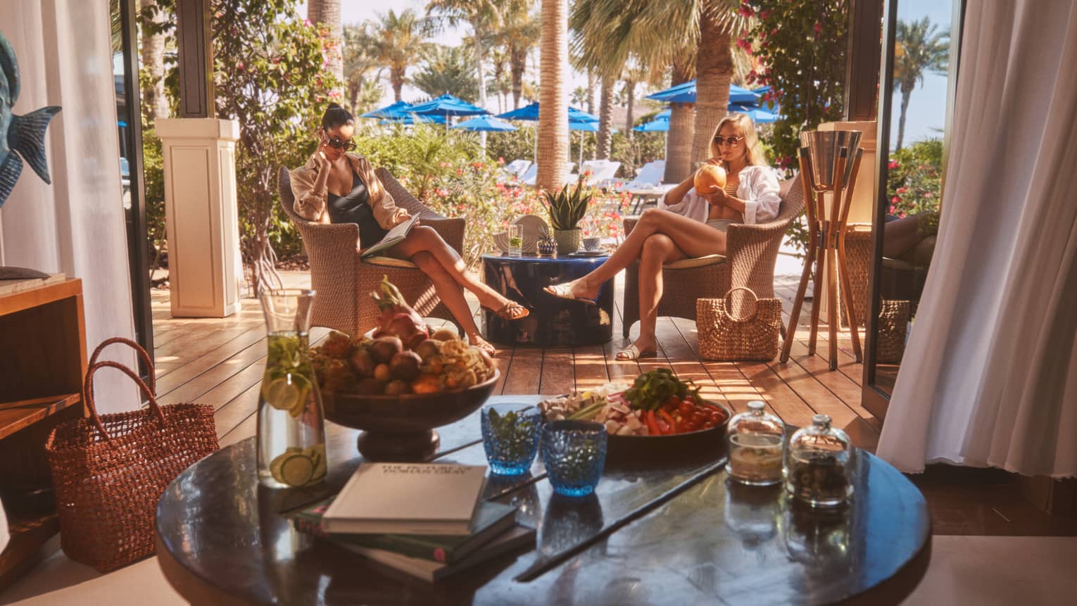 Two people sitting in rattan chairs with round table in forefront and blue pool umbrellas in backdrop