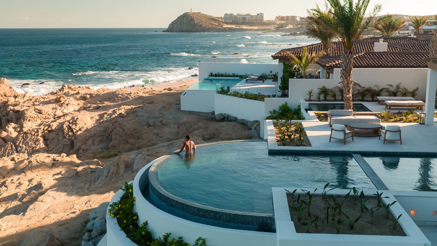 Oceanfront residence with an infinity pool, outdoor seating areas, palm trees and rocky coastline, featuring a person standing in the pool