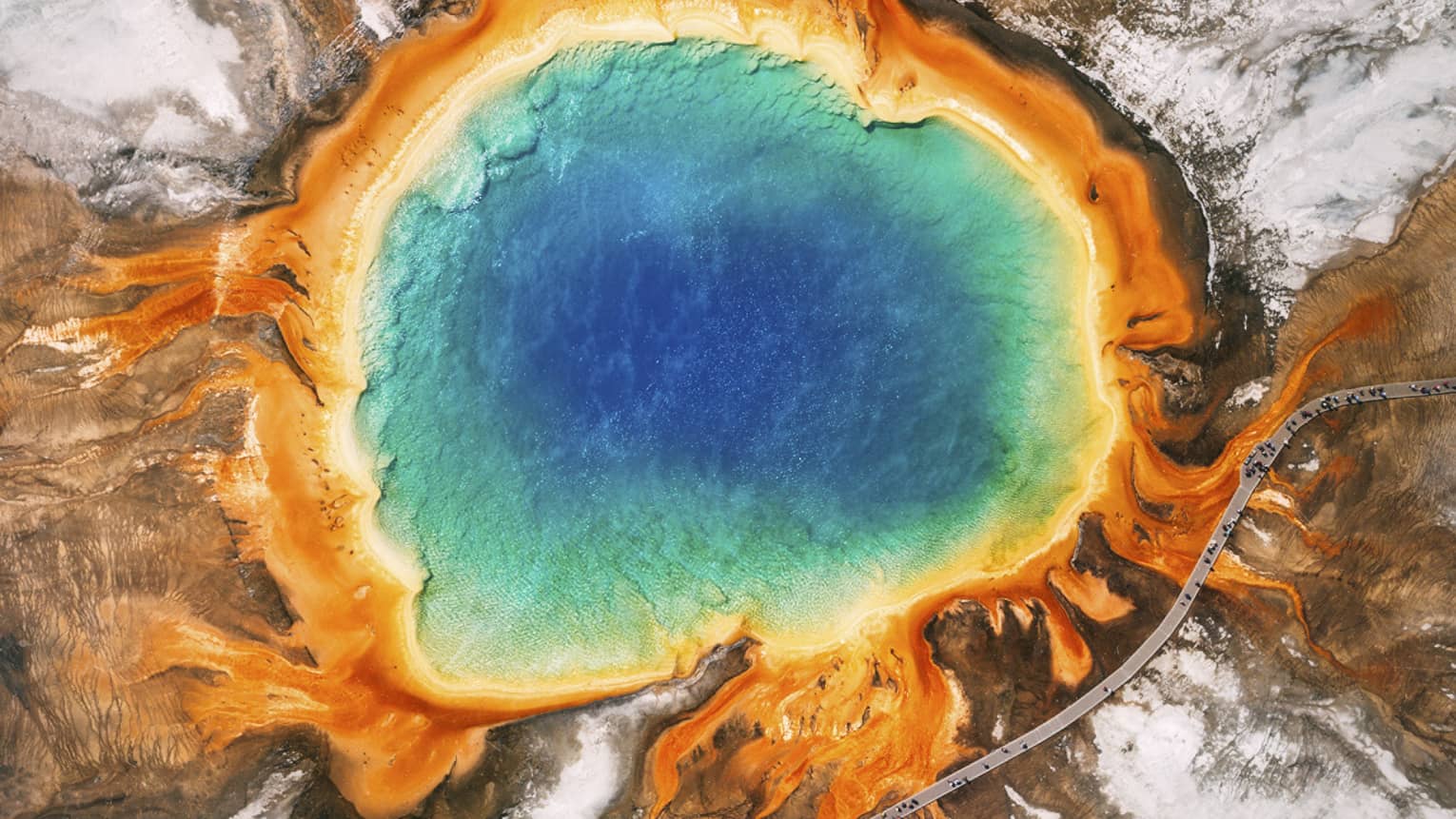 Aerial view of the Grand Prismatic Spring in Yellowstone, featuring vibrant blue, green, yellow and orange concentric rings, surrounded by rocky terrain