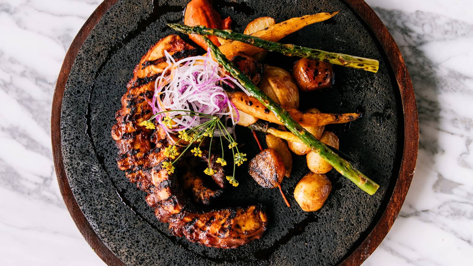 cooked octopus with potatoes and asparagus on a black plate.