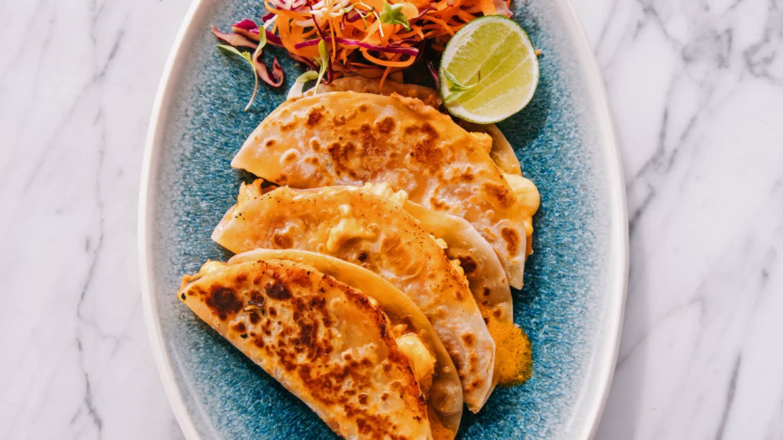 Three tacos and a brightly colored salad with a slice of lime on a blue plate create a visually appealing and appetizing dish.