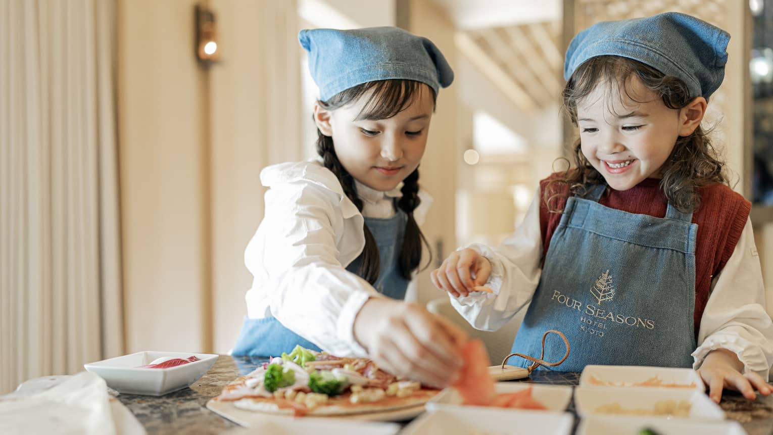 Two young girls in matching denim Four Seasons aprons and bandanas add toppings to their pizzas