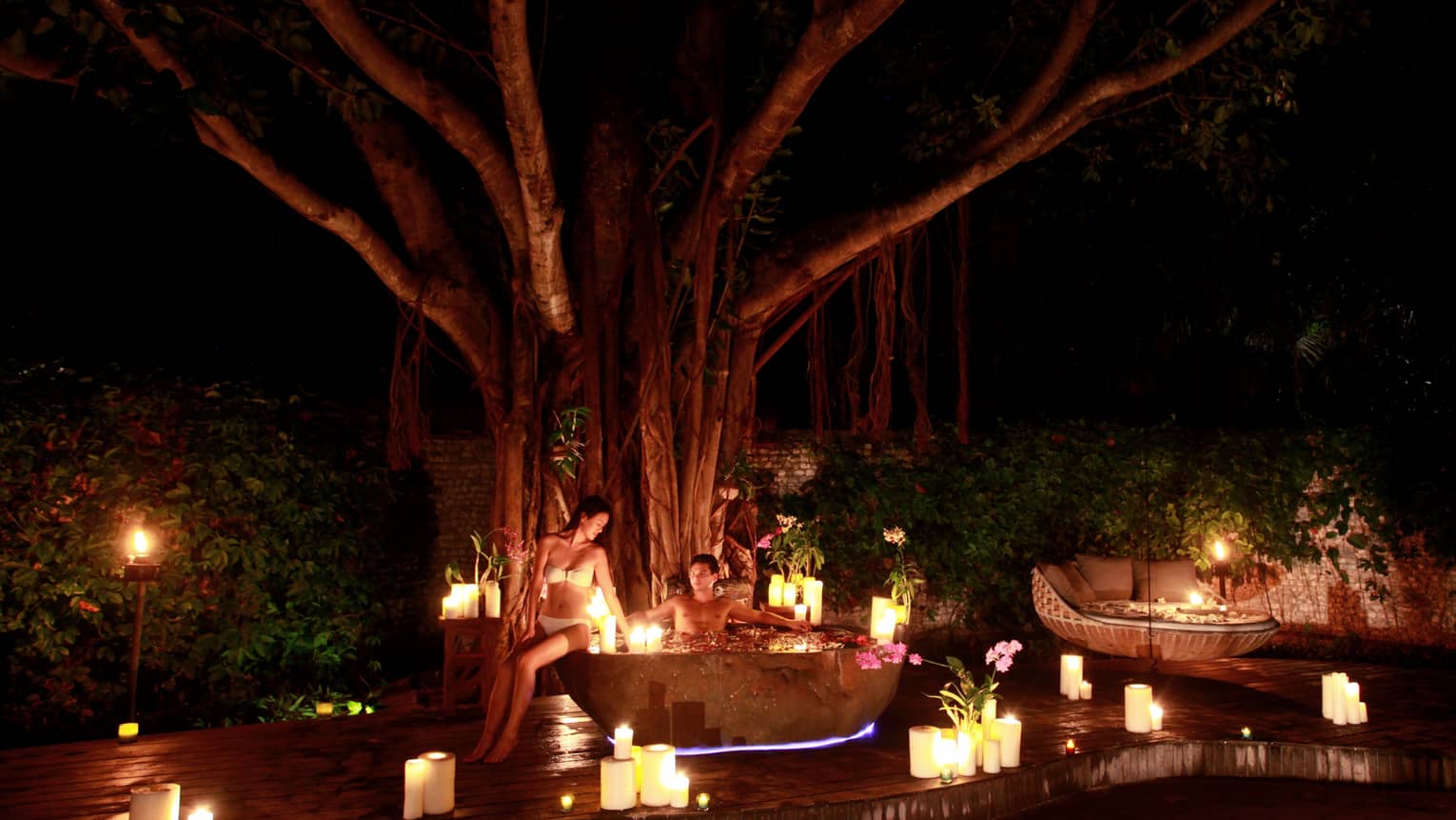 Man in outdoor whirlpool tub at night, woman in bikini on edge, surrounded by lit candles