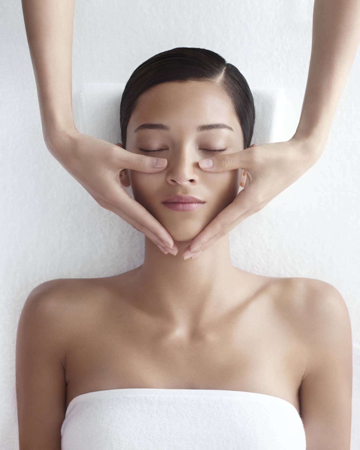 Woman in white bathrobe lies on massage table with eyes closed, masseuse rests hands on her cheeks