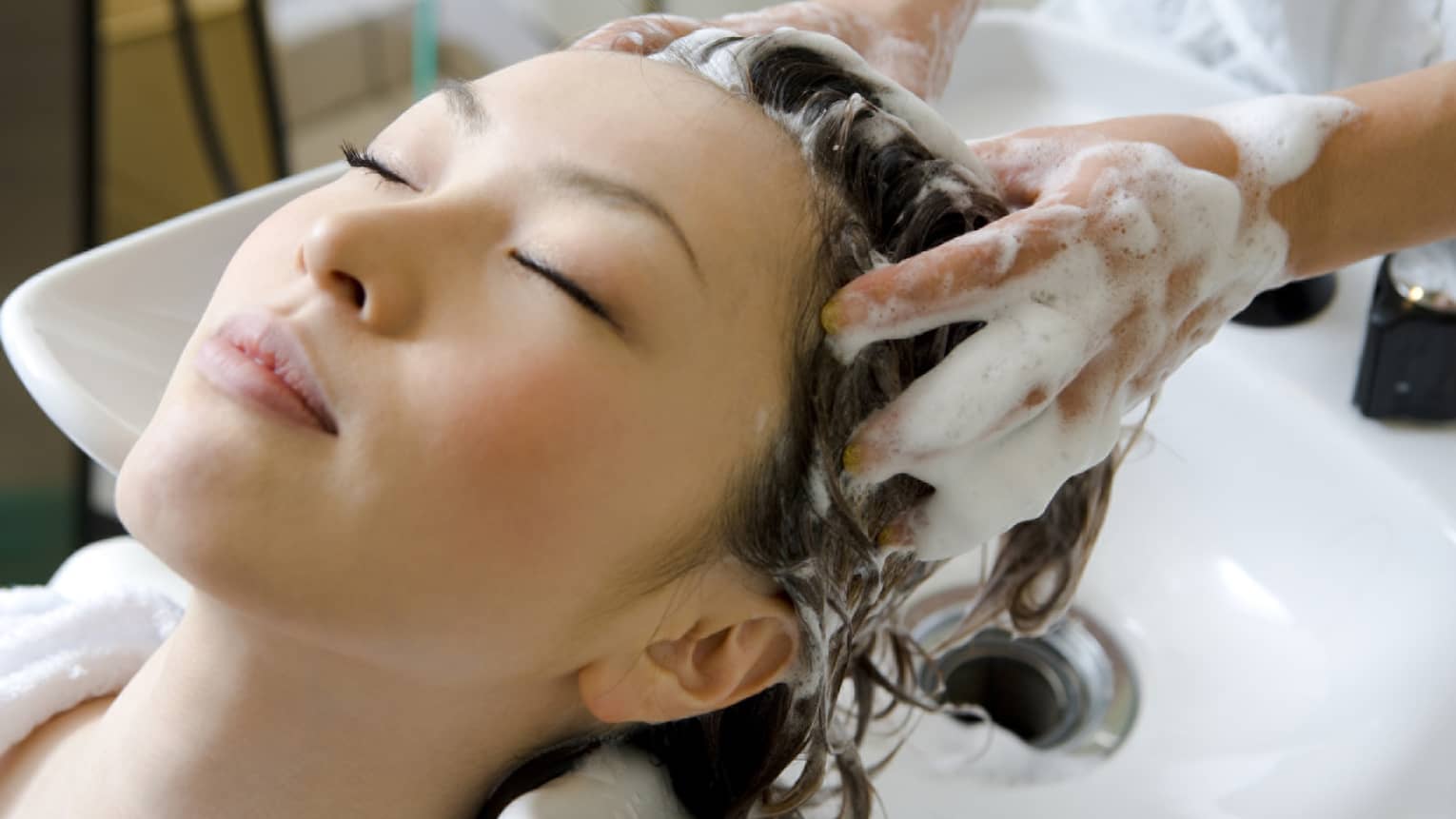 Woman closes eyes and leans back against sink as hands massage shampoo into her scalp