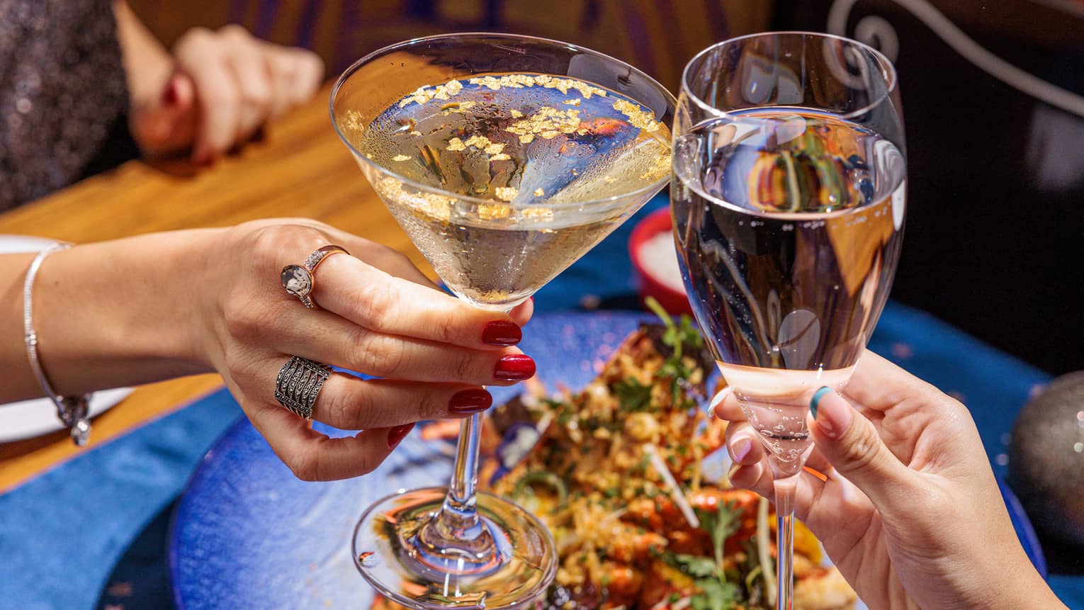 Two hands each holding stemmed glasses clink their glasses together over a bright blue plate filled with seafood