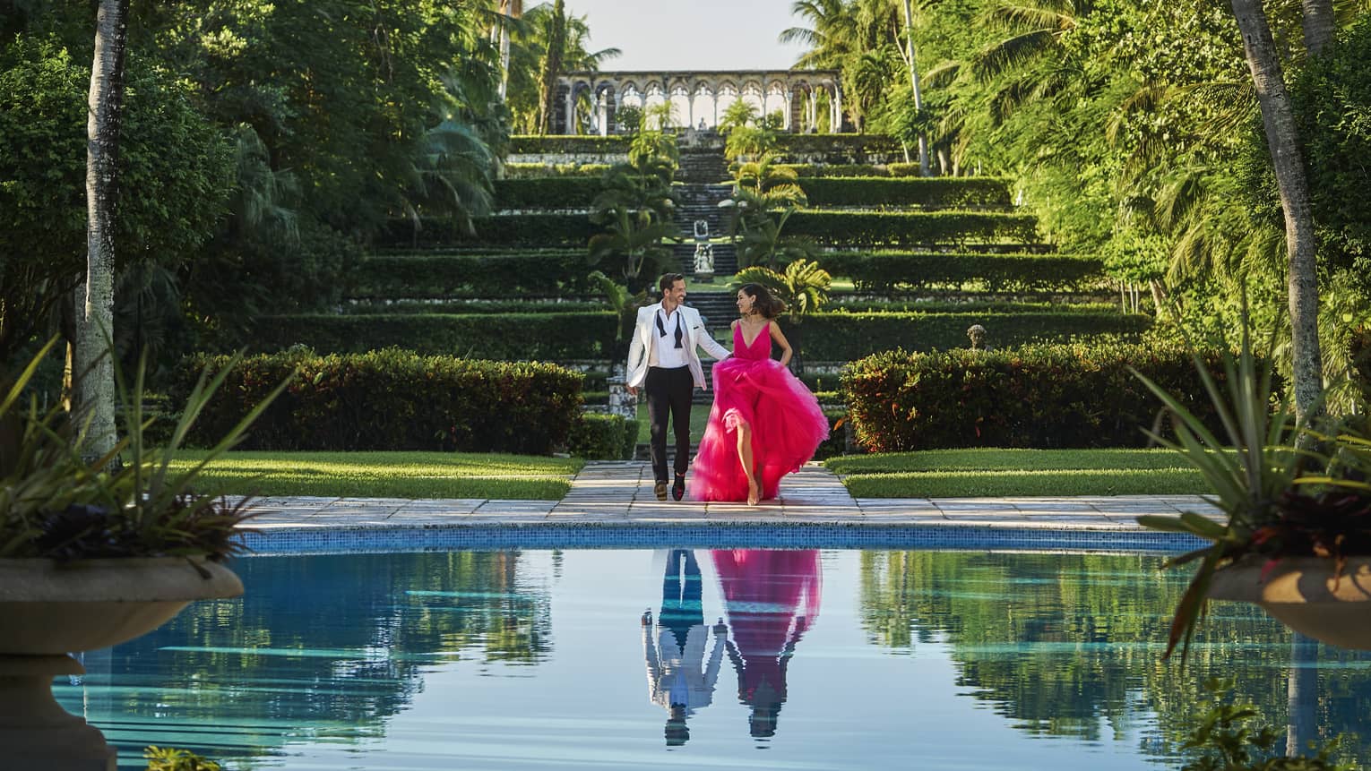 Man in suit and woman in pink dress standing near greenery and a lake.