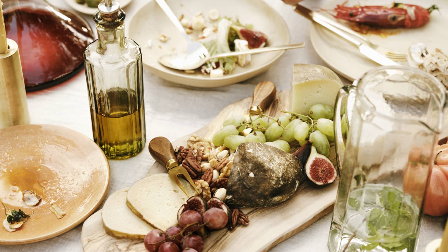 Close-up of a table laden with dishes of fresh figs, grapes, pecans, cheese, seafood, olive oil and a carafe of mint water.