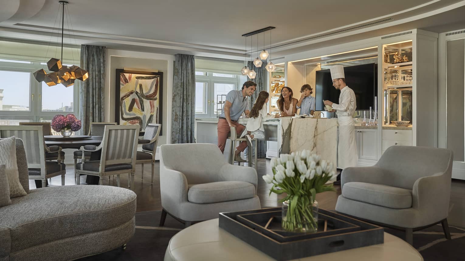 Luxury hotel suite with family interacting with a chef by a marble island, grey armchairs, tulips on ottoman, dining area, abstract art and city-view windows