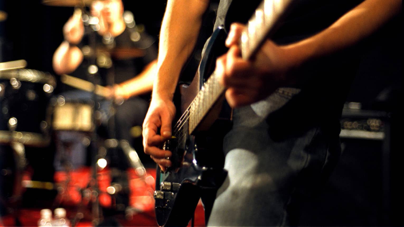 Close-up of musician's arms and hands playing an electric guitar with drumset in the background.
